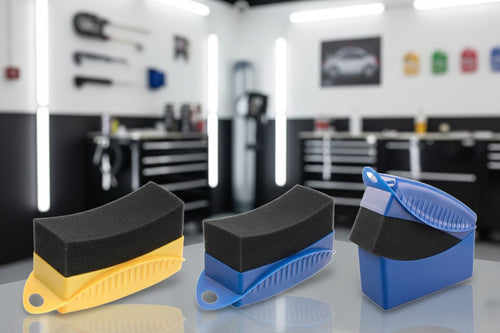 Car cleaning tools including a black sponge, blue brush, and yellow container on a car tire background.